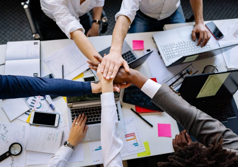 High-angle shot of diverse hands together over a desk with laptops and paperwork, symbolizing teamwork.